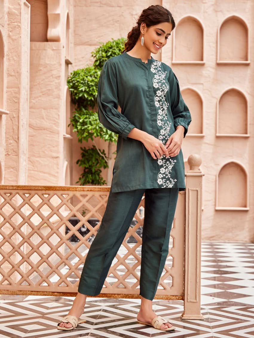 A woman standing in a beautifully decorated area wearing an embroidered green linen silk co-ord set with short sleeves and a partially elasticated waistband. She is also wearing light-colored floral embroidery on the chest area.