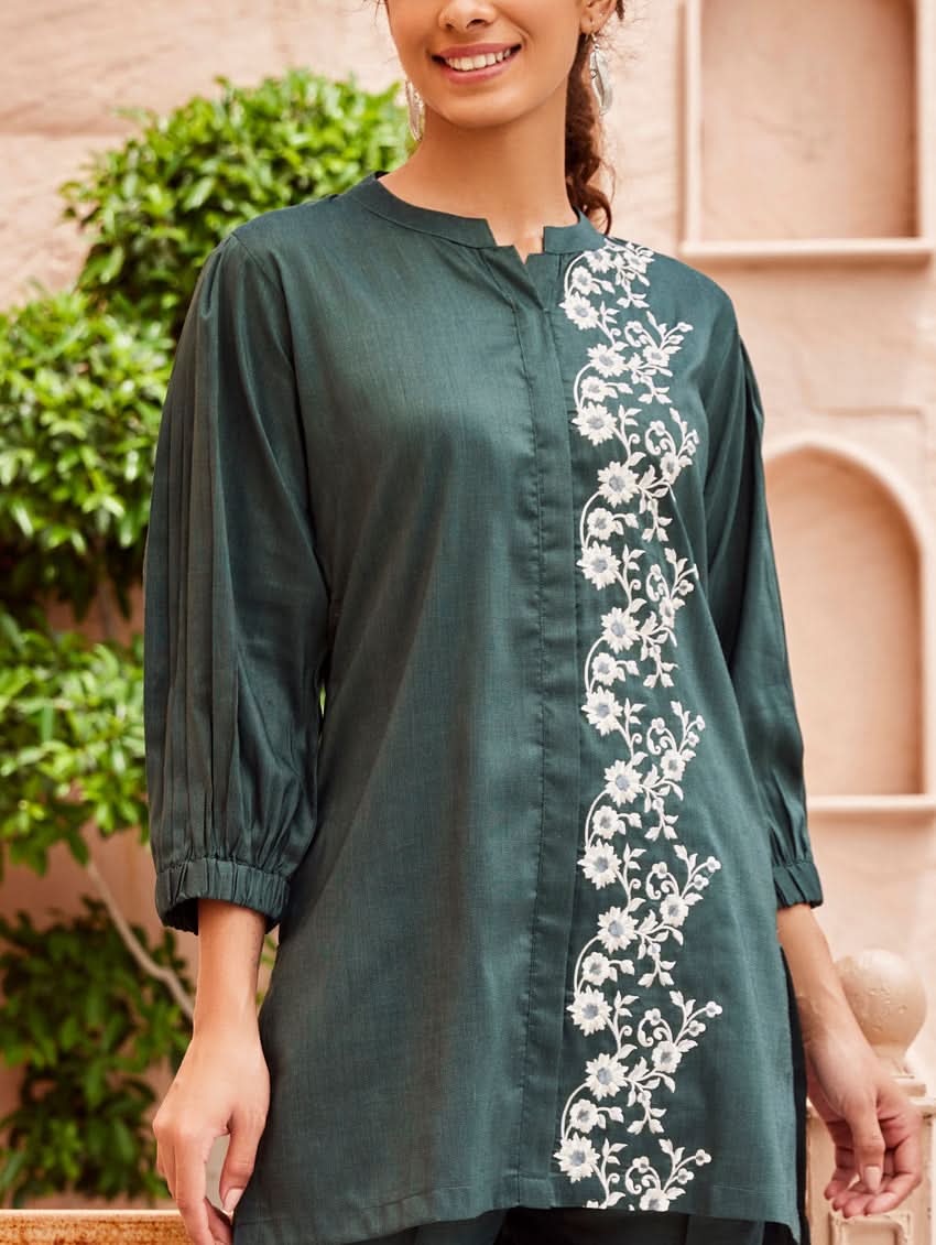 A woman standing in a beautifully decorated area wearing an embroidered green linen silk co-ord set with short sleeves and a partially elasticated waistband. She is also wearing light-colored floral embroidery on the chest area.
