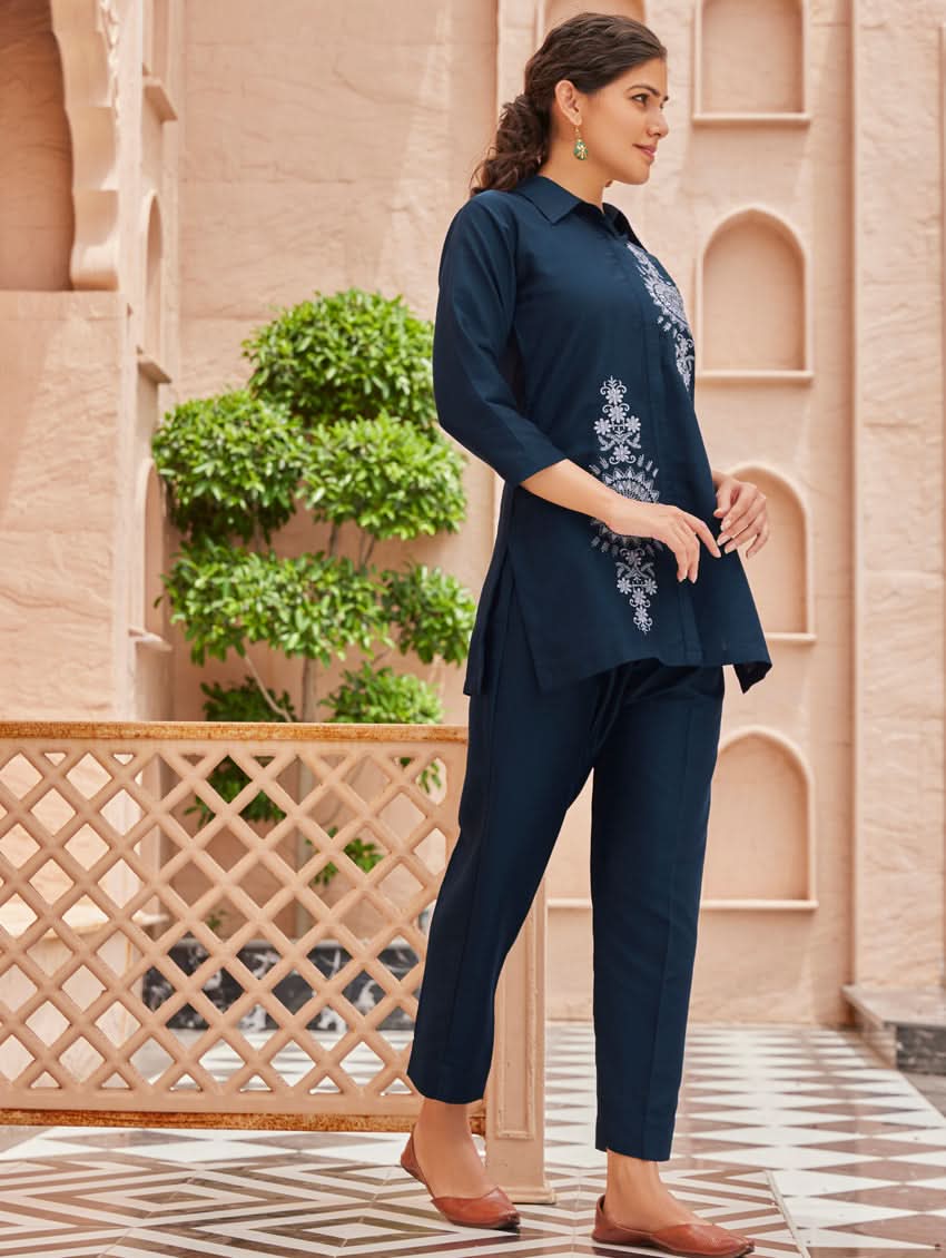 A woman standing outdoors wearing an embroidered viscose rayon co-ord set with a rever neckline, three-quarter sleeves, and pockets on either side.