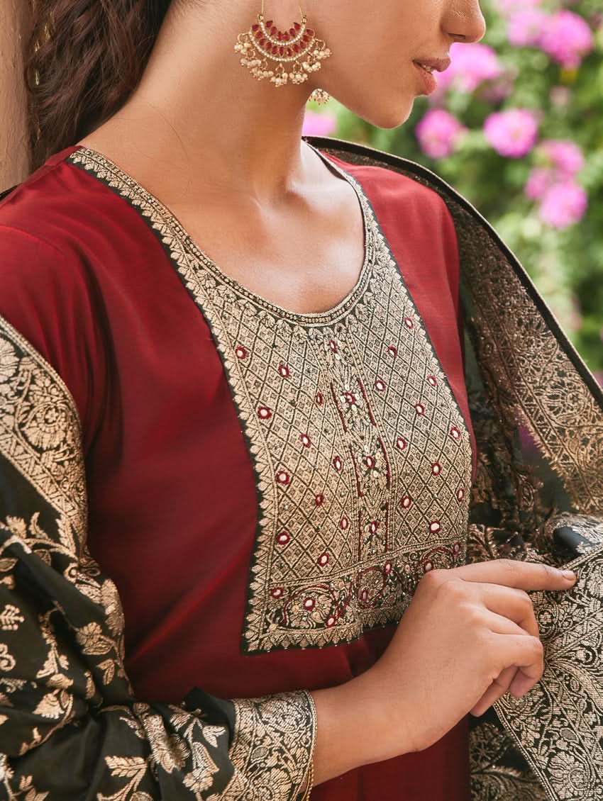 A woman wearing a hand-embroidered butter silk suit set with a jacquard weave silk dupatta in a red and dark color scheme, standing near a decorative entrance.