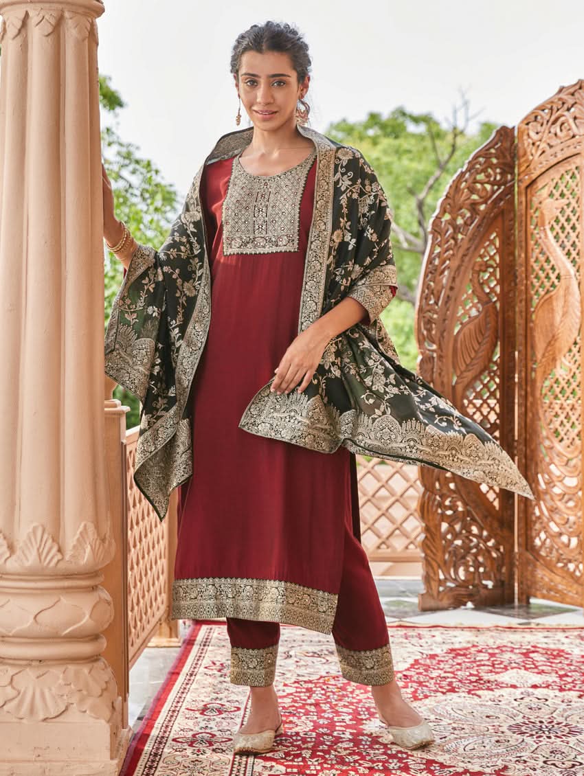 A woman wearing a hand-embroidered butter silk suit set with a jacquard weave silk dupatta in a red and dark color scheme, standing near a decorative entrance.