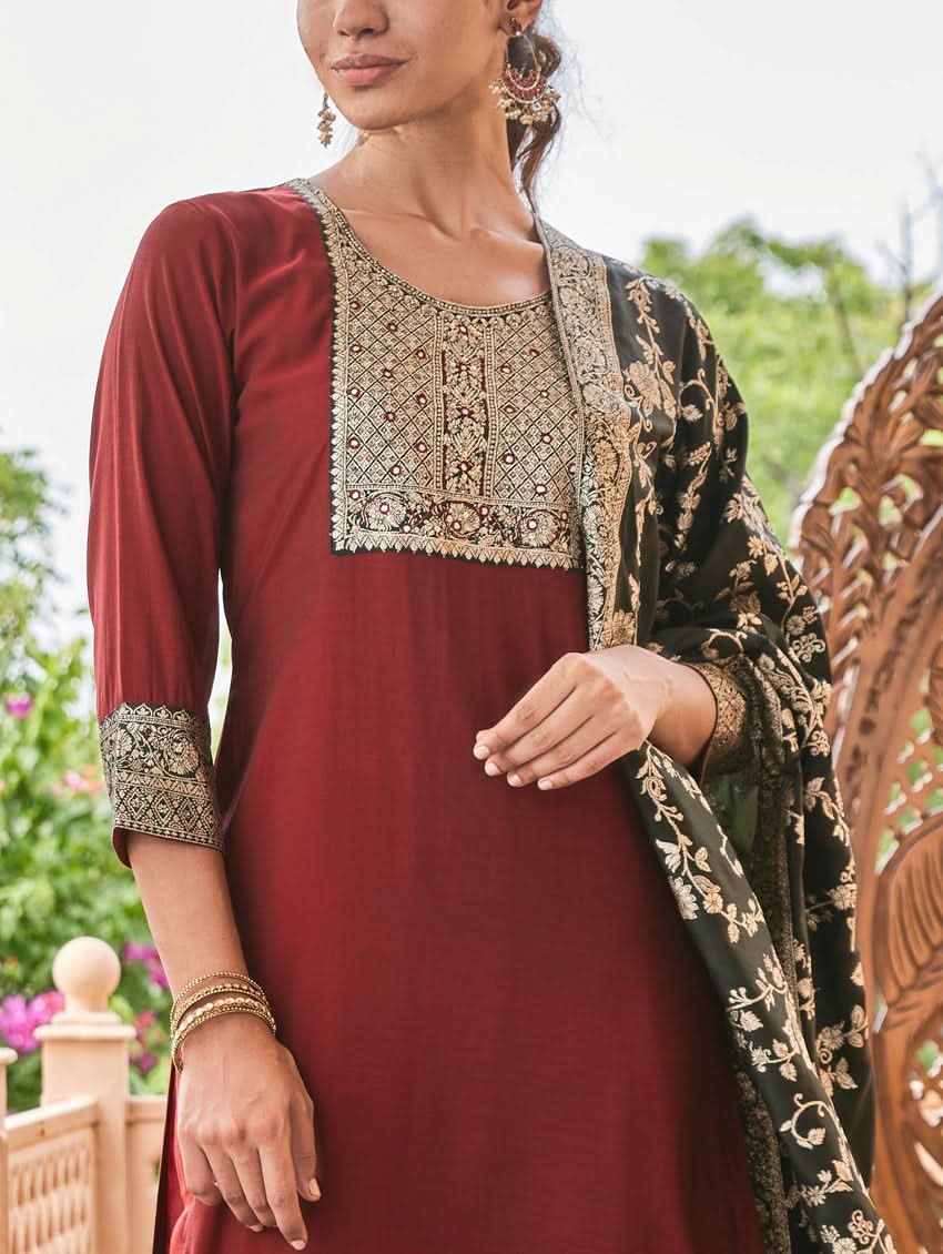 A woman wearing a hand-embroidered butter silk suit set with a jacquard weave silk dupatta in a red and dark color scheme, standing near a decorative entrance.