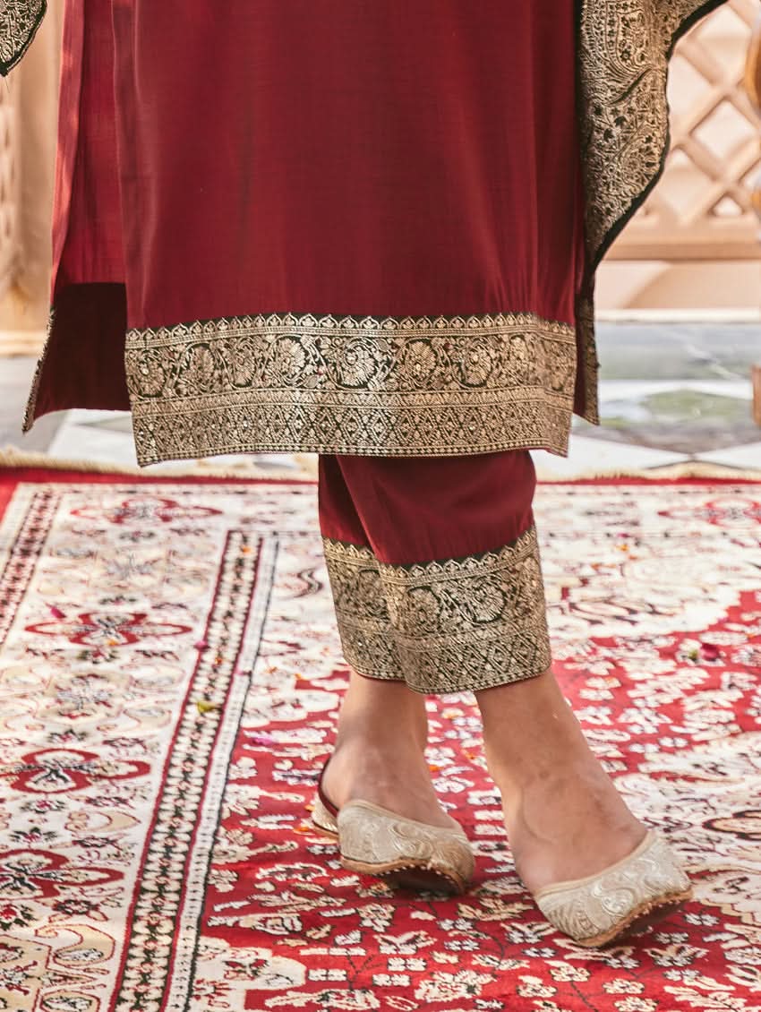 A woman wearing a hand-embroidered butter silk suit set with a jacquard weave silk dupatta in a red and dark color scheme, standing near a decorative entrance.