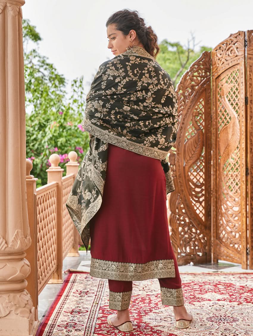 A woman wearing a hand-embroidered butter silk suit set with a jacquard weave silk dupatta in a red and dark color scheme, standing near a decorative entrance.