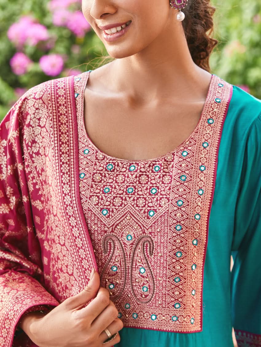 A woman standing in a traditional setting, wearing a blue silk suit with embroidered details and a dupatta with solid print and jacquard dupatta.