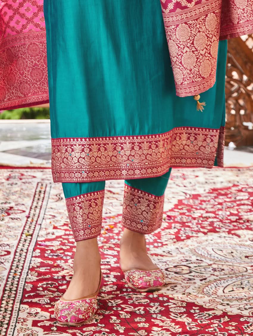 A woman standing in a traditional setting, wearing a blue silk suit with embroidered details and a dupatta with solid print and jacquard dupatta.