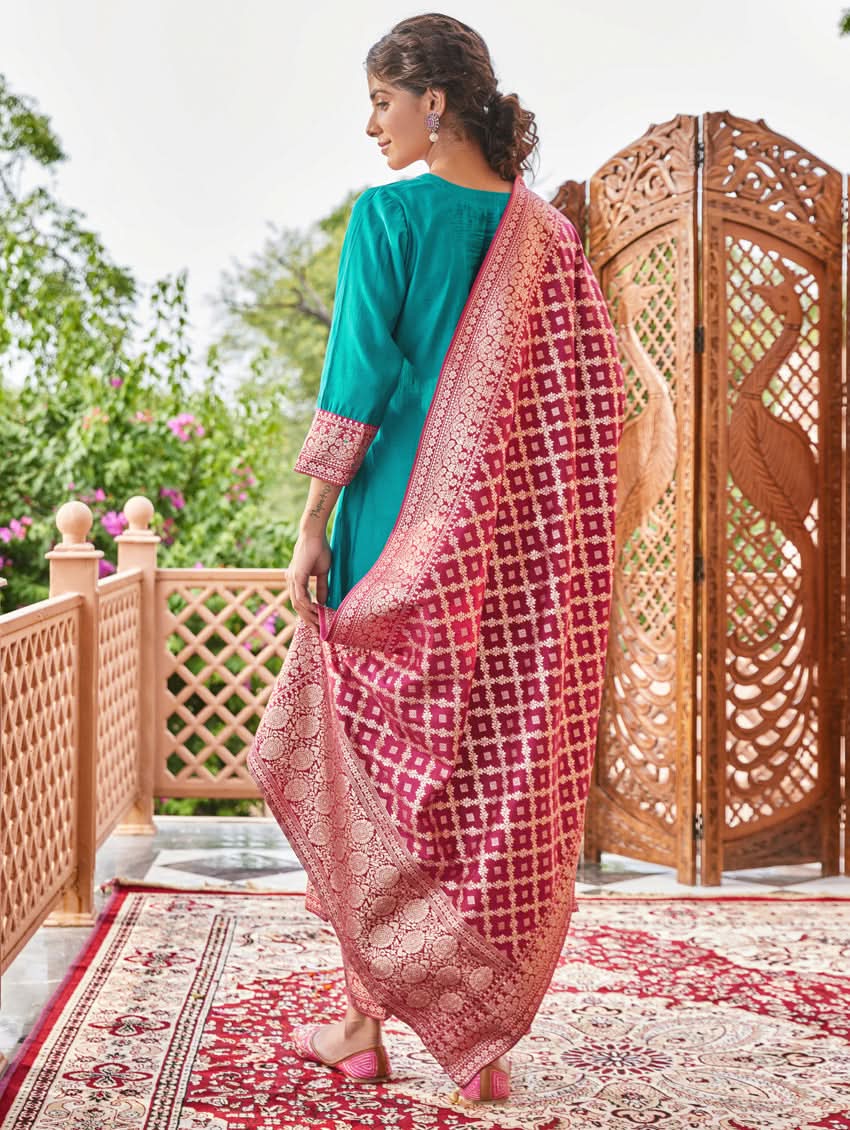A woman standing in a traditional setting, wearing a blue silk suit with embroidered details and a dupatta with solid print and jacquard dupatta.