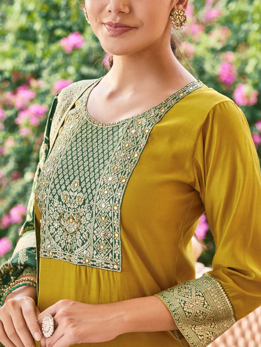 A woman wearing a traditional green and gold silk suit set with hand embroidery, featuring a kurta with round neckline and three-fourth sleeves, and pants with a partially elasticated waistband. The set is displayed outdoors with a floral backdrop.