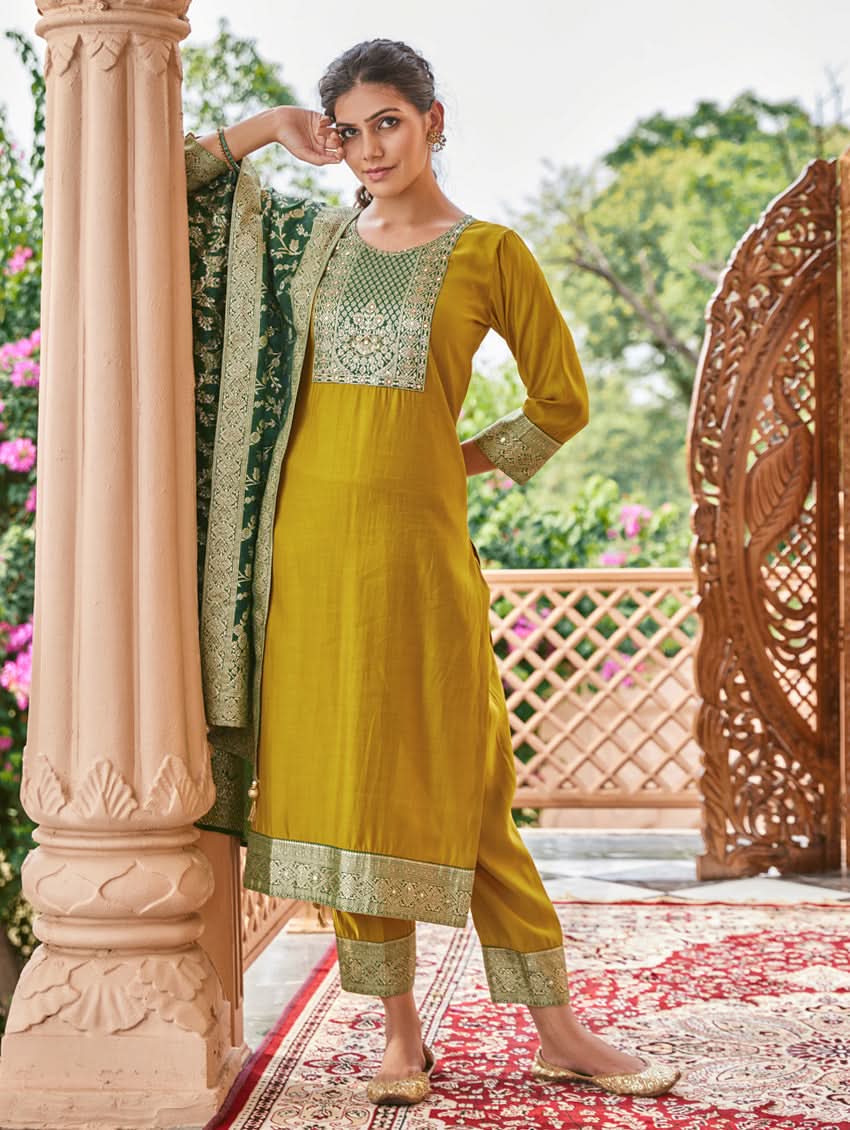 A woman wearing a traditional green and gold silk suit set with hand embroidery, featuring a kurta with round neckline and three-fourth sleeves, and pants with a partially elasticated waistband. The set is displayed outdoors with a floral backdrop.