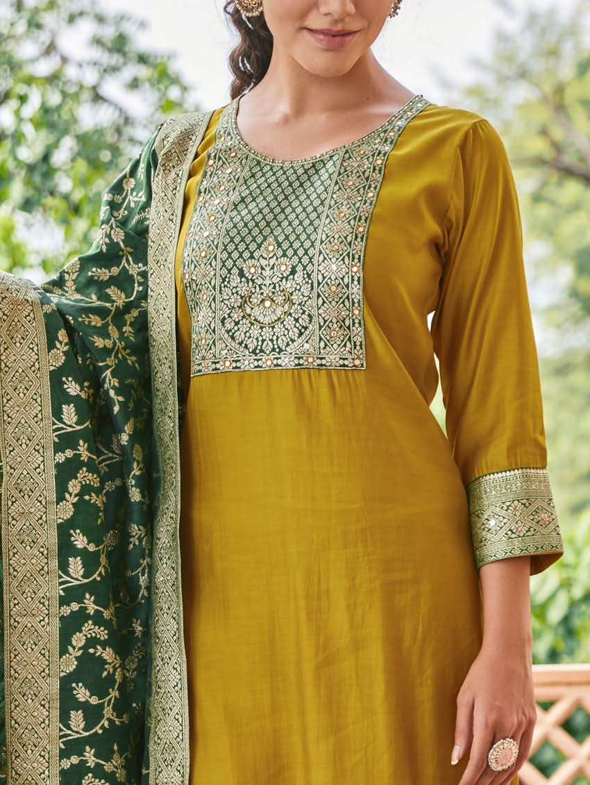 A woman wearing a traditional green and gold silk suit set with hand embroidery, featuring a kurta with round neckline and three-fourth sleeves, and pants with a partially elasticated waistband. The set is displayed outdoors with a floral backdrop.