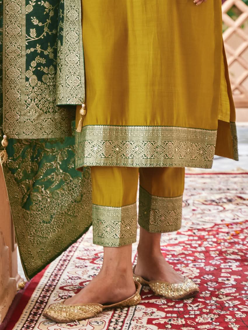 A woman wearing a traditional green and gold silk suit set with hand embroidery, featuring a kurta with round neckline and three-fourth sleeves, and pants with a partially elasticated waistband. The set is displayed outdoors with a floral backdrop.