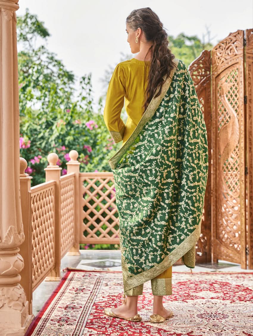 A woman wearing a traditional green and gold silk suit set with hand embroidery, featuring a kurta with round neckline and three-fourth sleeves, and pants with a partially elasticated waistband. The set is displayed outdoors with a floral backdrop.
