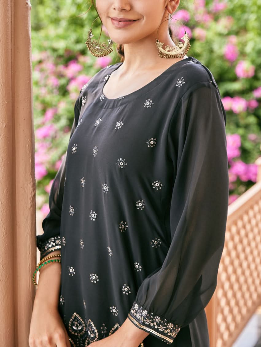 A woman standing outdoors wearing an embroidered georgette co-ord set with a round neckline, three-quarter sleeves, and pockets on either side. The set is predominantly black with embellishments in a sequin or computerized thread on the kurta.