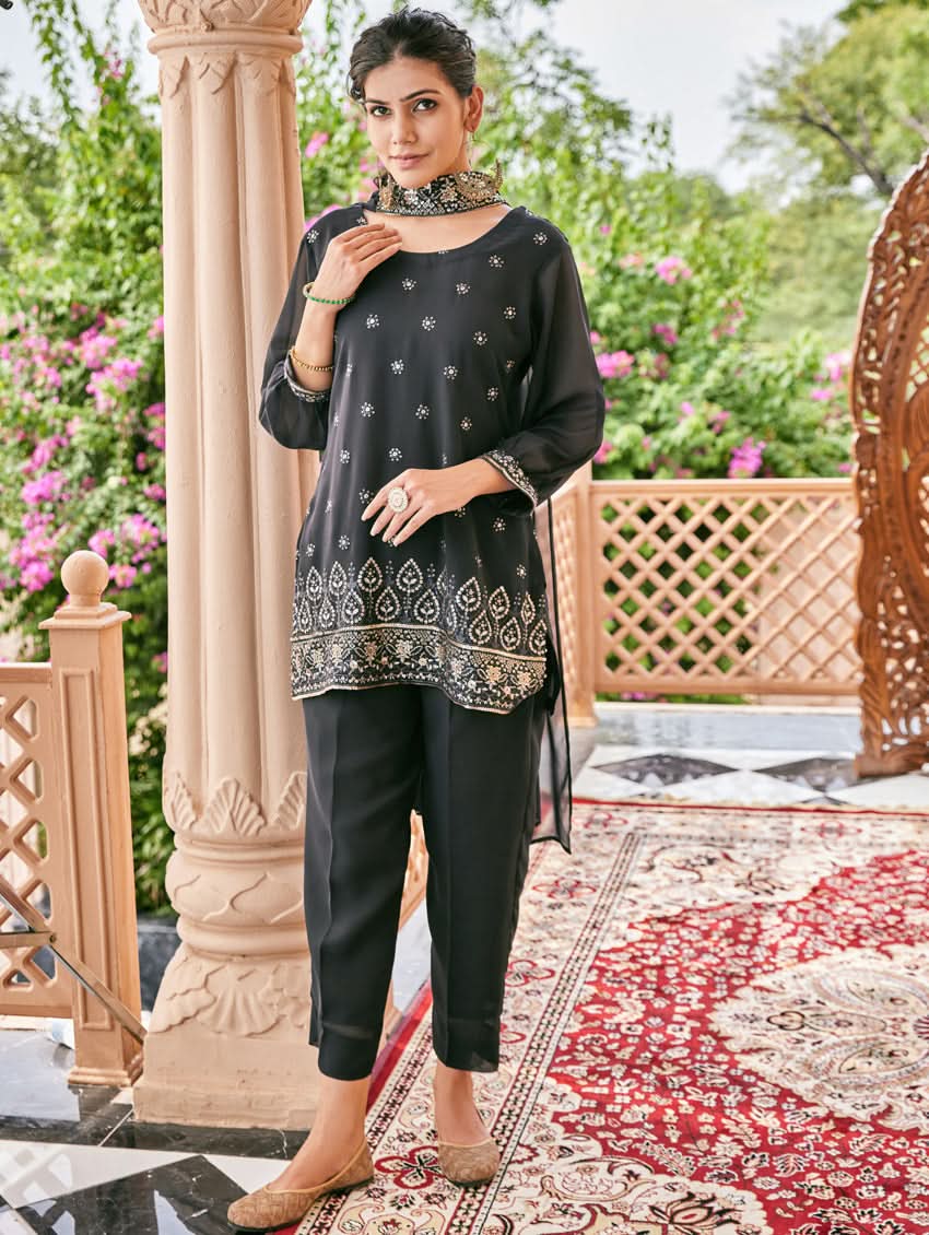 A woman standing outdoors wearing an embroidered georgette co-ord set with a round neckline, three-quarter sleeves, and pockets on either side. The set is predominantly black with embellishments in a sequin or computerized thread on the kurta.