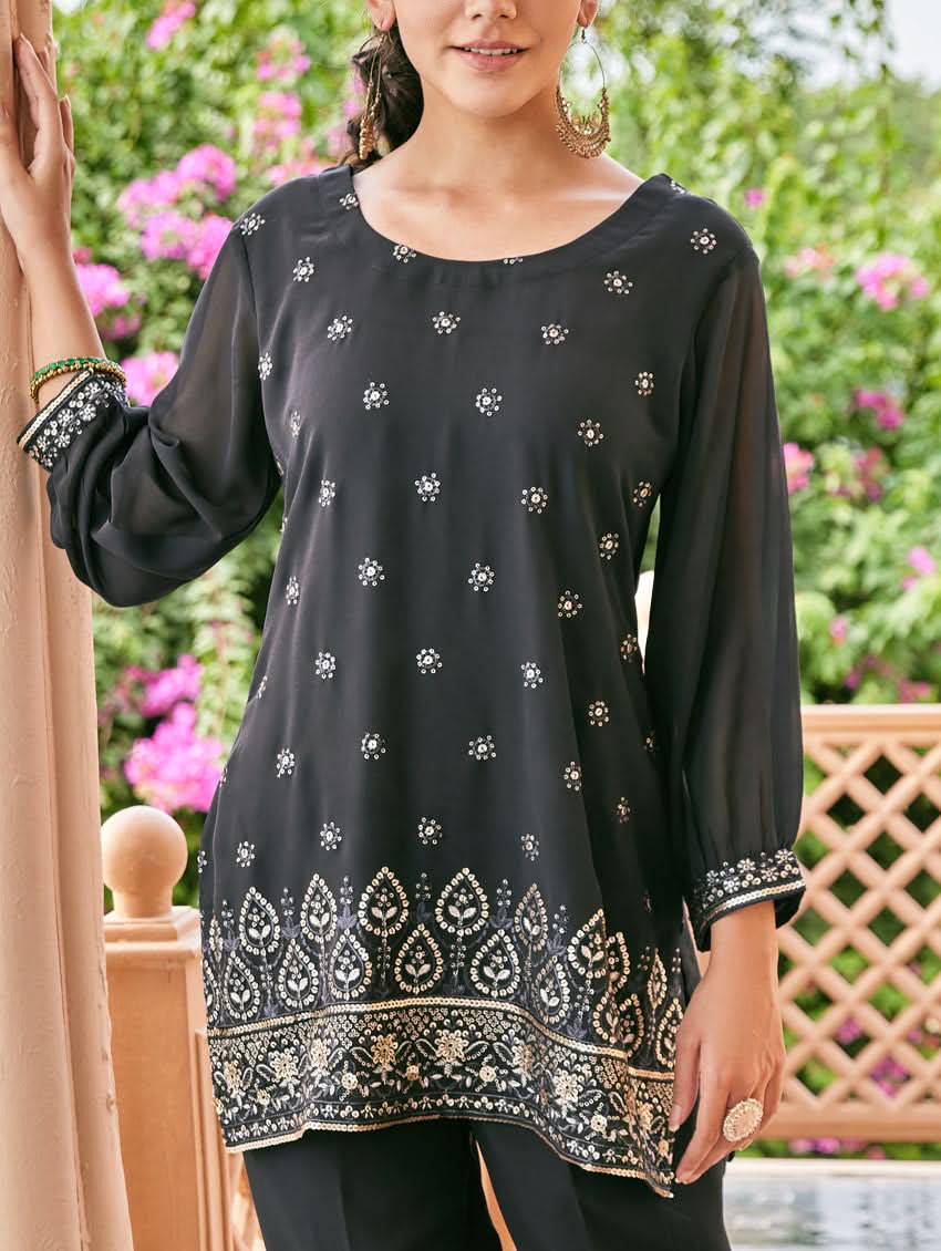 A woman standing outdoors wearing an embroidered georgette co-ord set with a round neckline, three-quarter sleeves, and pockets on either side. The set is predominantly black with embellishments in a sequin or computerized thread on the kurta.