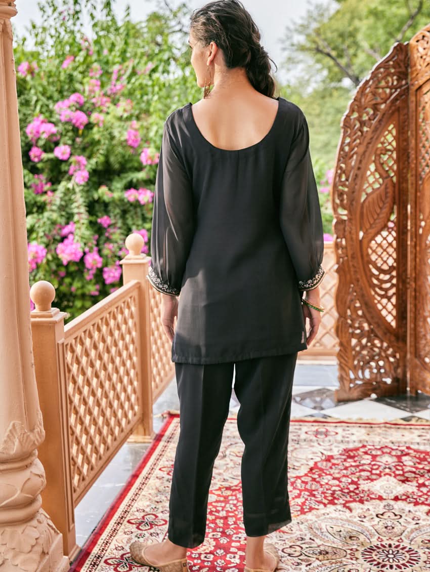 A woman standing outdoors wearing an embroidered georgette co-ord set with a round neckline, three-quarter sleeves, and pockets on either side. The set is predominantly black with embellishments in a sequin or computerized thread on the kurta.