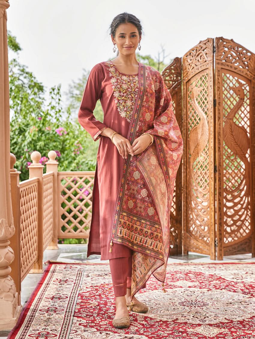 A woman standing in an elegant pose wearing a red silk suit set with floral patterns on the dupatta, straight pants, and a kurta with pearl work.
