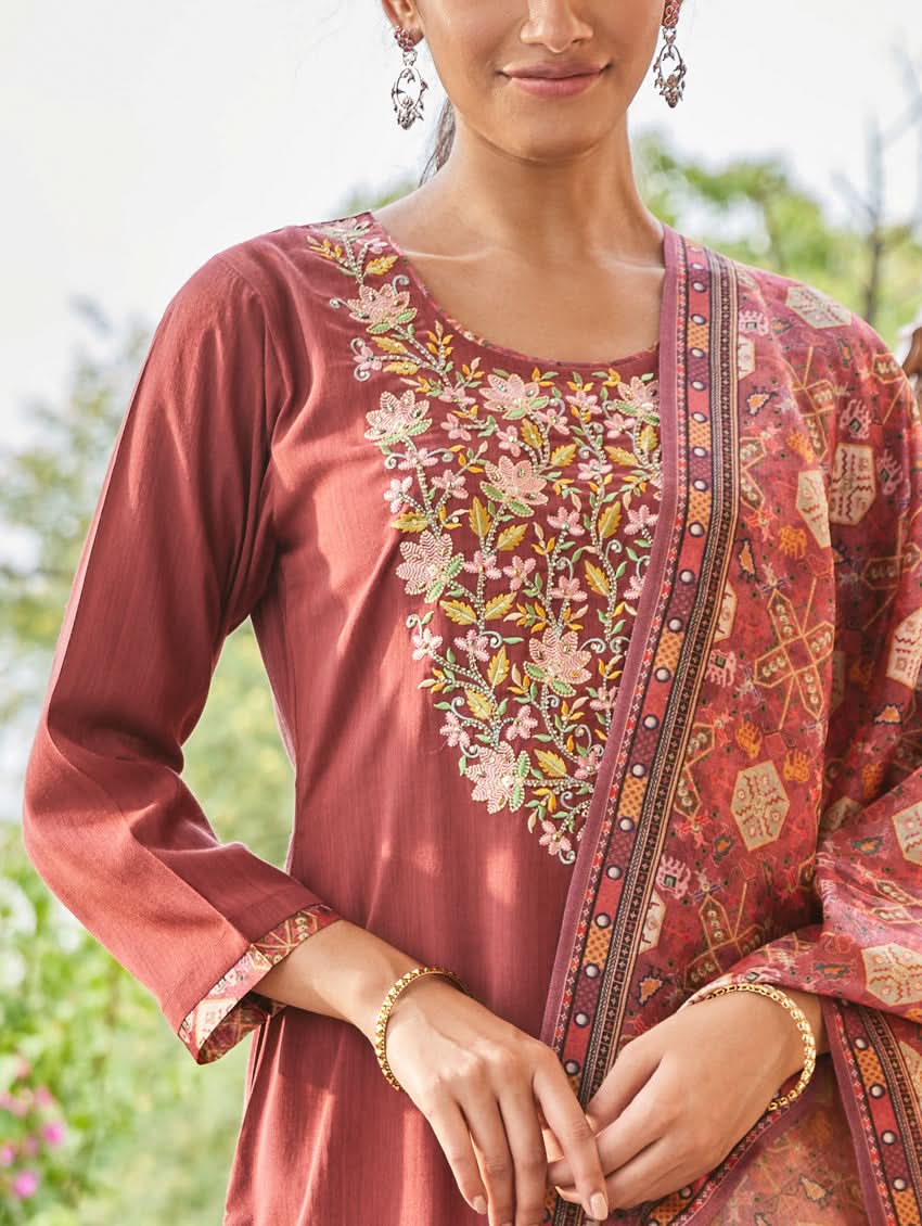 A woman standing in an elegant pose wearing a red silk suit set with floral patterns on the dupatta, straight pants, and a kurta with pearl work.