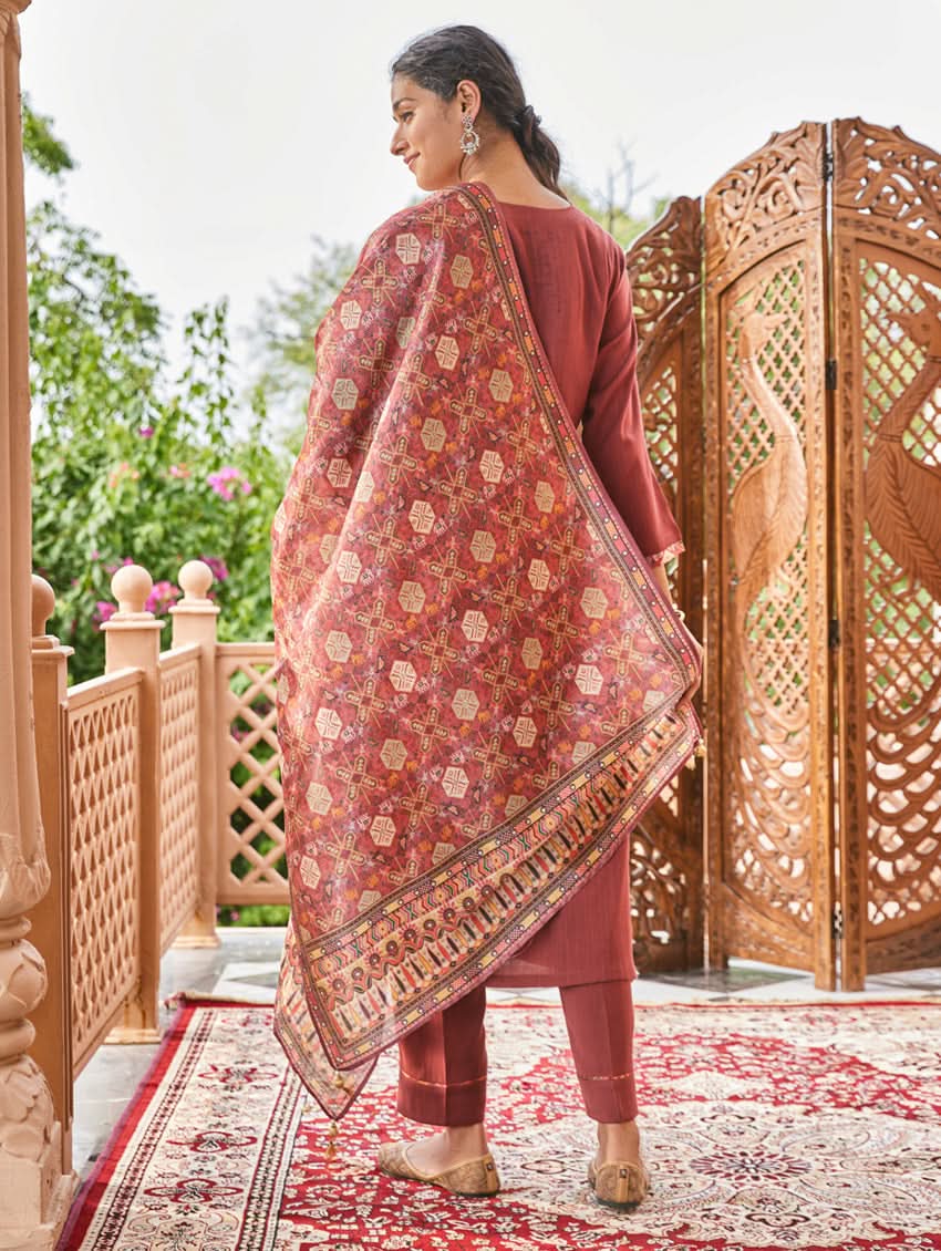 A woman standing in an elegant pose wearing a red silk suit set with floral patterns on the dupatta, straight pants, and a kurta with pearl work.