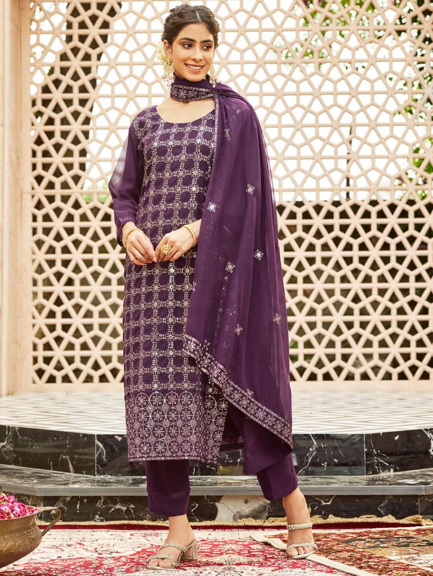 A woman wearing a traditional purple georgette suit with embroidery and a dupatta, standing in a courtyard with a decorative wall in the background.