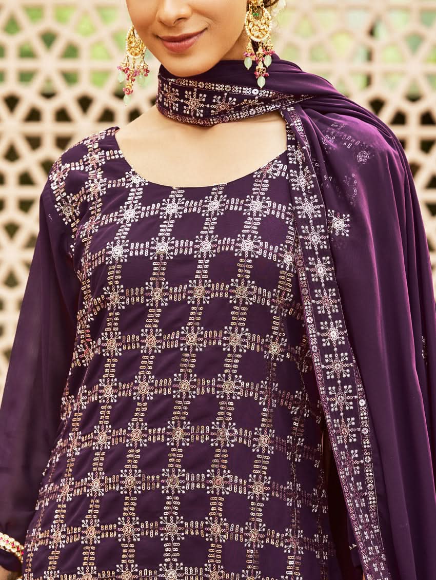 A woman wearing a traditional purple georgette suit with embroidery and a dupatta, standing in a courtyard with a decorative wall in the background.