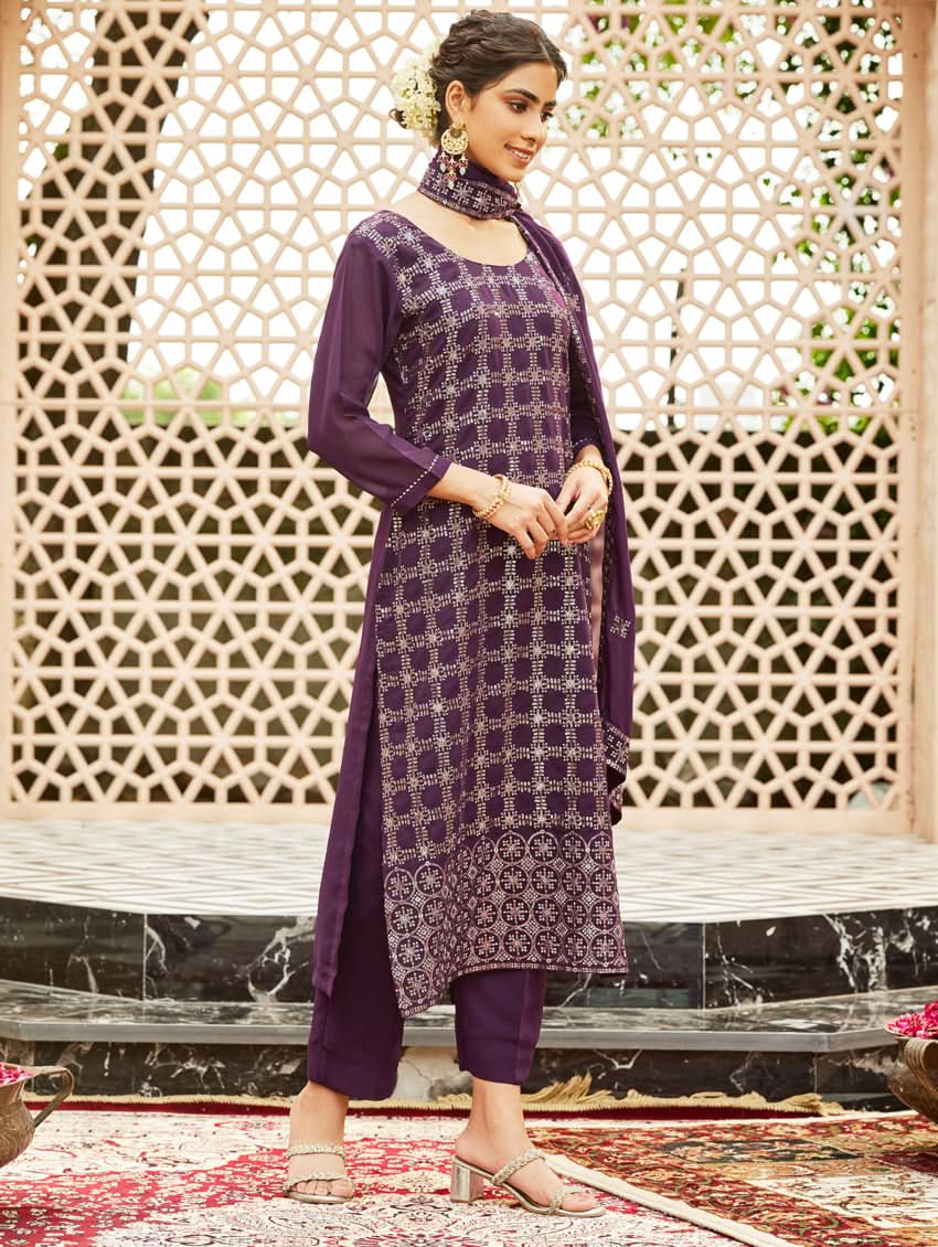 A woman wearing a traditional purple georgette suit with embroidery and a dupatta, standing in a courtyard with a decorative wall in the background.