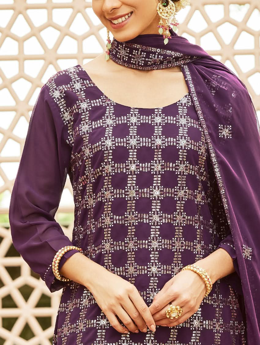 A woman wearing a traditional purple georgette suit with embroidery and a dupatta, standing in a courtyard with a decorative wall in the background.