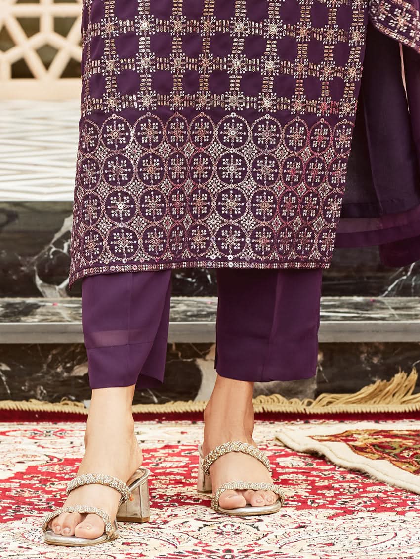 A woman wearing a traditional purple georgette suit with embroidery and a dupatta, standing in a courtyard with a decorative wall in the background.