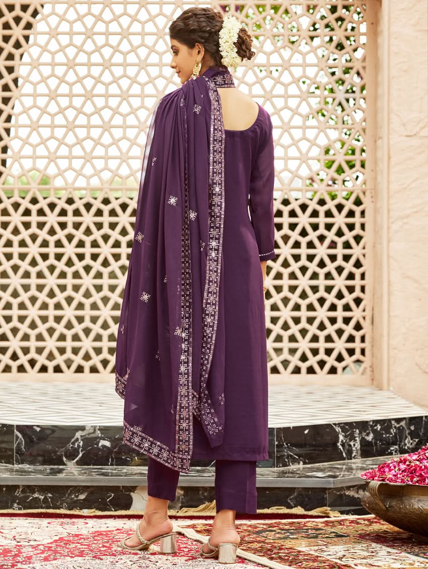 A woman wearing a traditional purple georgette suit with embroidery and a dupatta, standing in a courtyard with a decorative wall in the background.