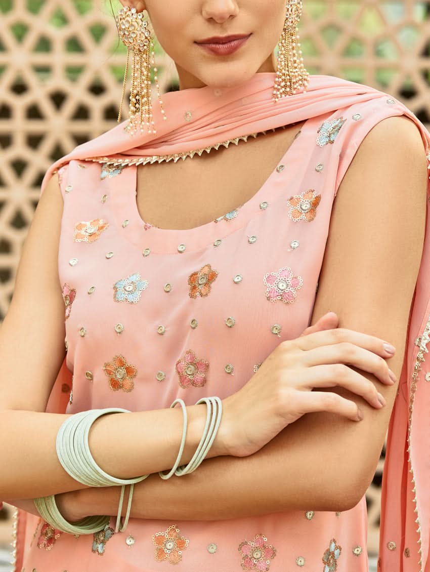 A model wearing a pink embroidered georgette sharara suit set with a round neckline and sleeveless design, featuring sequin work and a dupatta.