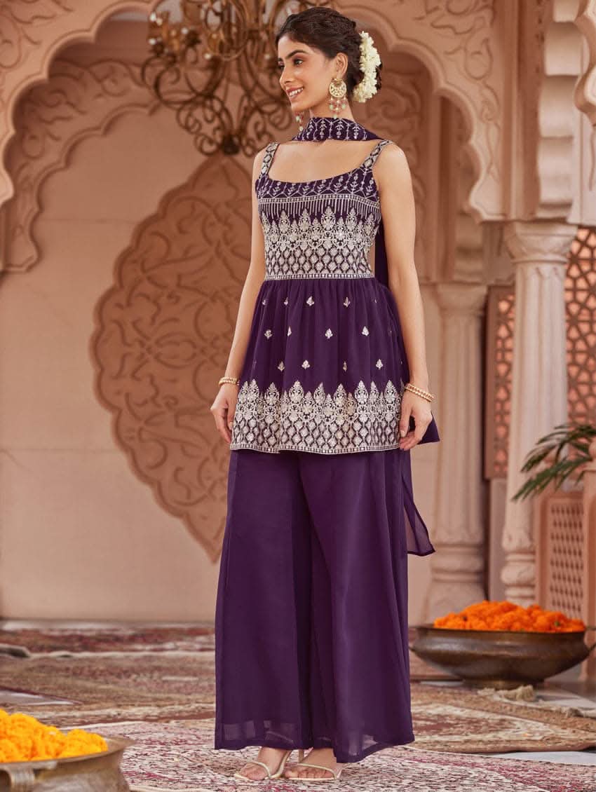 A woman wearing a purple peplum style suit set with embroidered kurta and sharara, standing in a beautifully decorated area.
