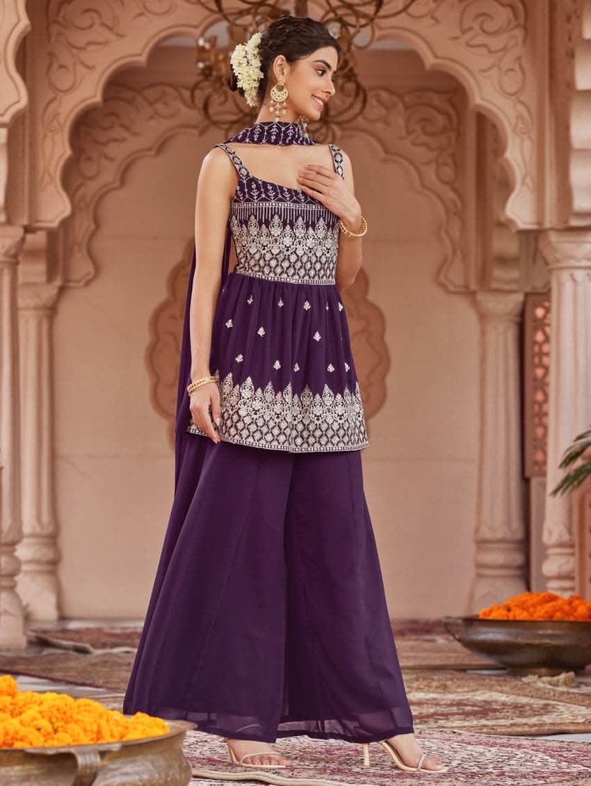 A woman wearing a purple peplum style suit set with embroidered kurta and sharara, standing in a beautifully decorated area.
