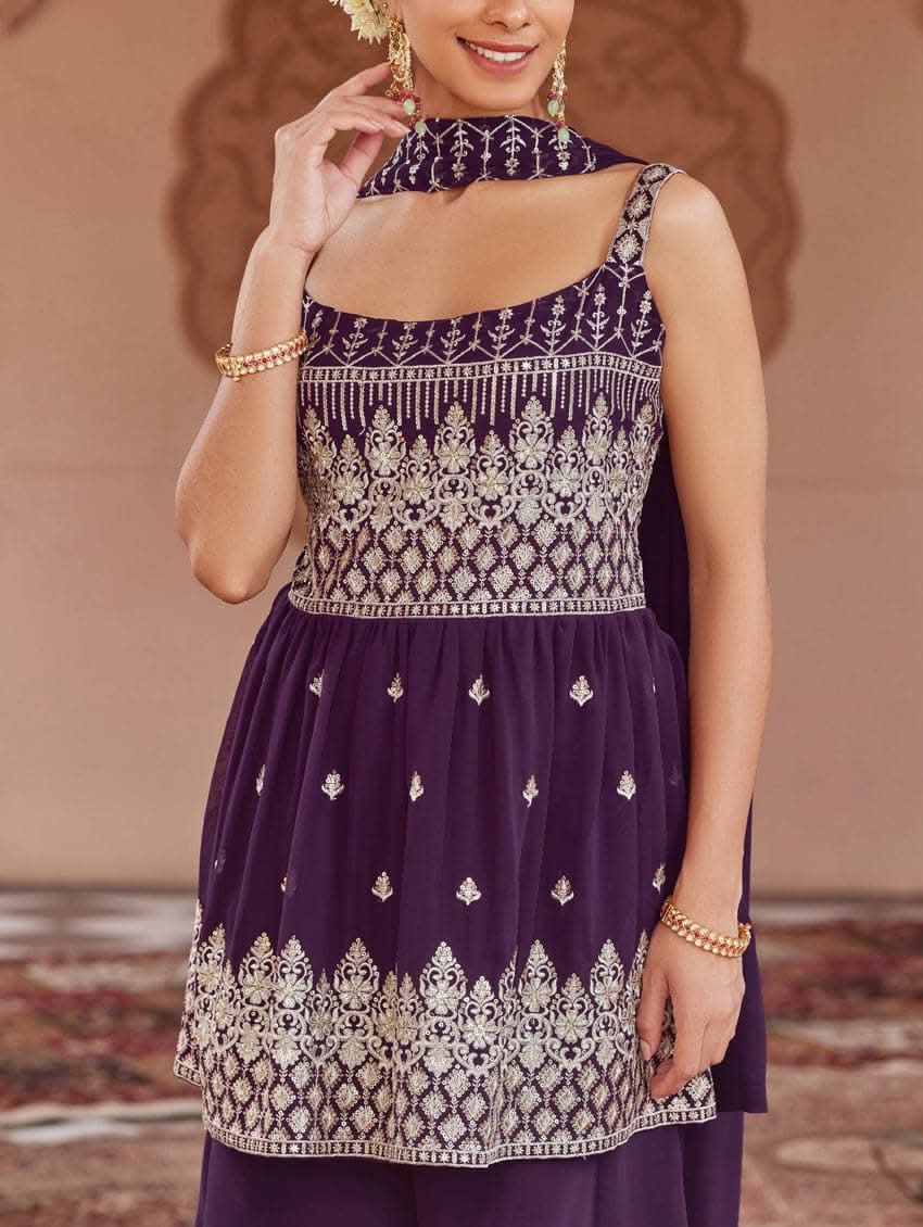 A woman wearing a purple peplum style suit set with embroidered kurta and sharara, standing in a beautifully decorated area.