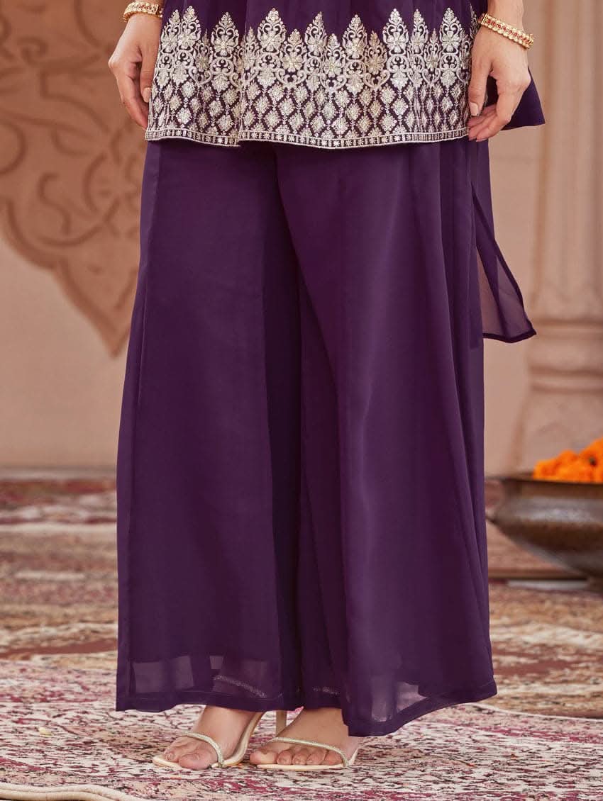 A woman wearing a purple peplum style suit set with embroidered kurta and sharara, standing in a beautifully decorated area.