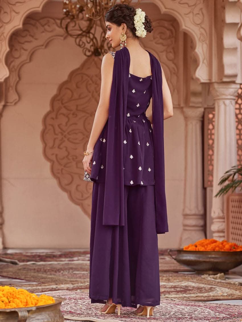 A woman wearing a purple peplum style suit set with embroidered kurta and sharara, standing in a beautifully decorated area.
