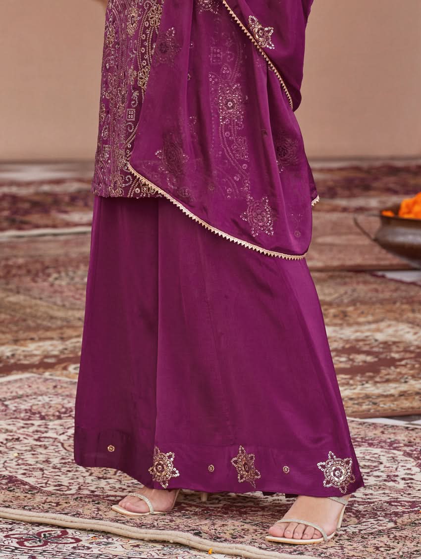 A woman wearing a traditional purple Georgette suit set with embroidered sequins, consisting of a kurta with round neckline and sleeveless design, and a sharara with an elasticated waistband. The set is complemented by a dupatta.