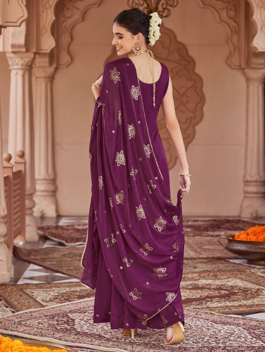 A woman wearing a traditional purple Georgette suit set with embroidered sequins, consisting of a kurta with round neckline and sleeveless design, and a sharara with an elasticated waistband. The set is complemented by a dupatta.