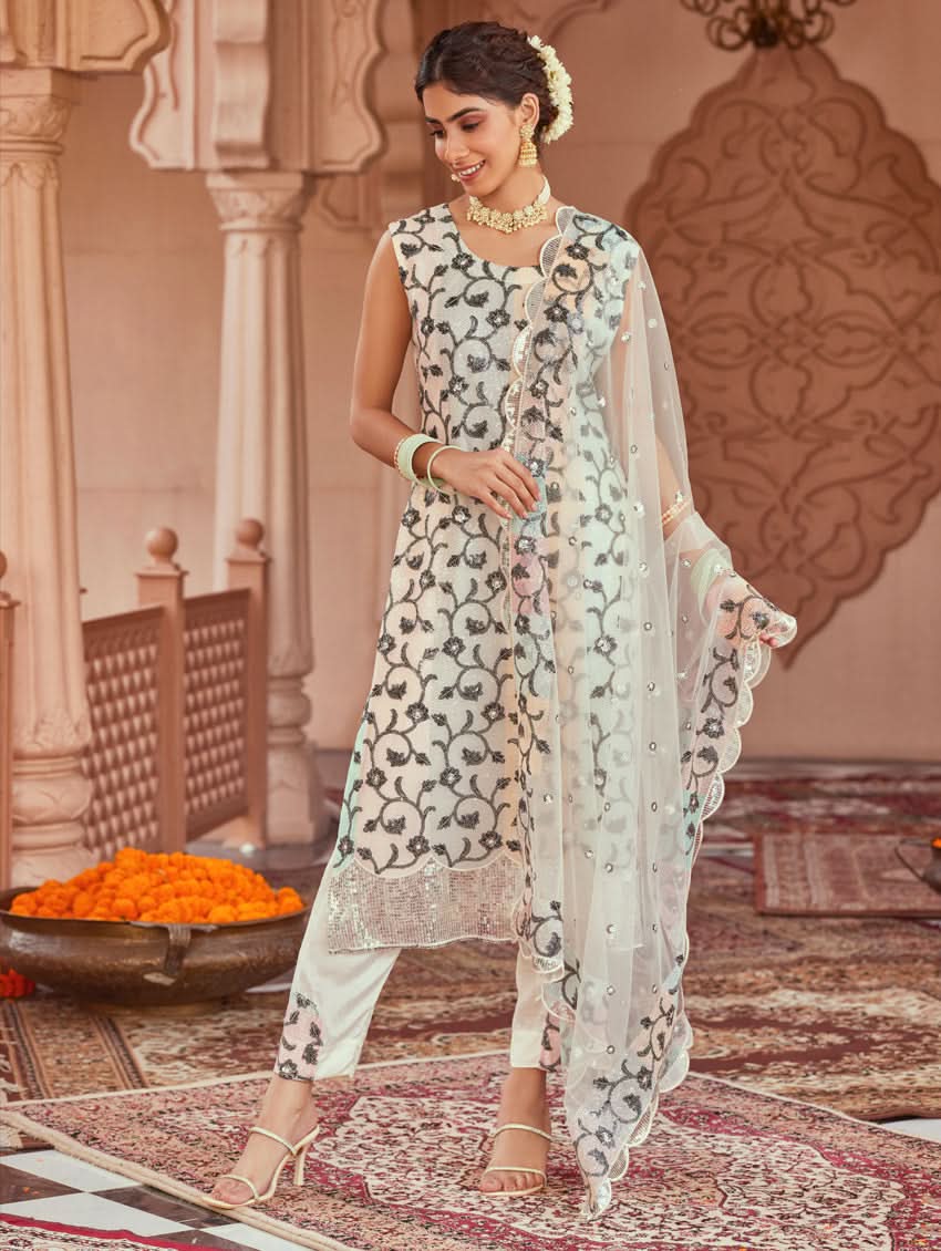A woman wearing a white net suit set with embroidery and sequence work, consisting of a kurta, pants, and dupatta, standing in a festive setting.