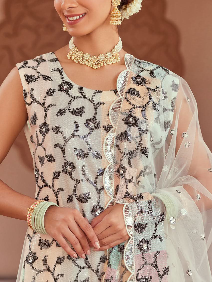 A woman wearing a white net suit set with embroidery and sequence work, consisting of a kurta, pants, and dupatta, standing in a festive setting.