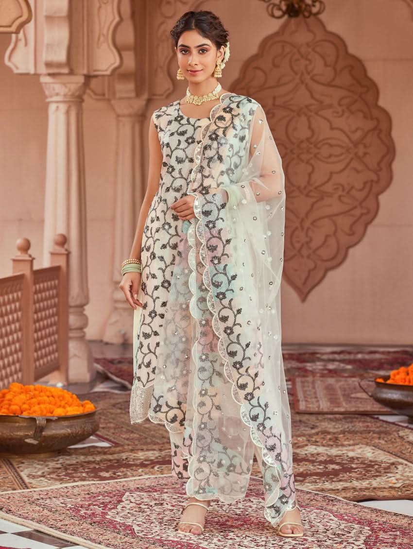 A woman wearing a white net suit set with embroidery and sequence work, consisting of a kurta, pants, and dupatta, standing in a festive setting.