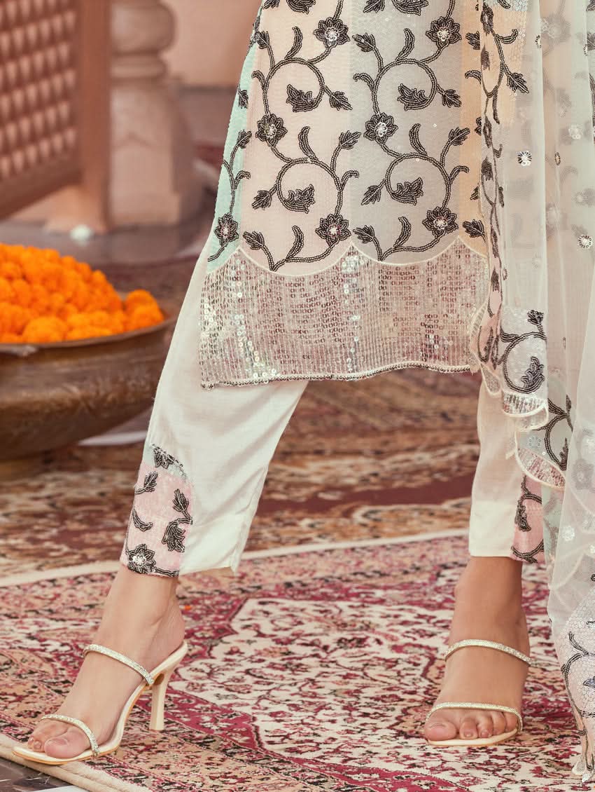 A woman wearing a white net suit set with embroidery and sequence work, consisting of a kurta, pants, and dupatta, standing in a festive setting.