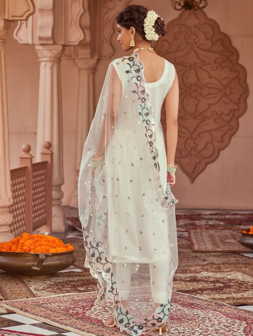 A woman wearing a white net suit set with embroidery and sequence work, consisting of a kurta, pants, and dupatta, standing in a festive setting.