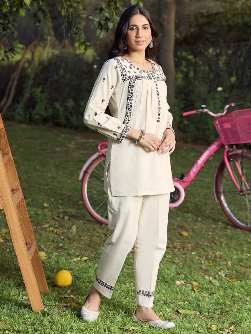 A woman standing outdoors wearing a beige embroidered viscose flex co-ord set with short kurta and pants, next to a pink bicycle.