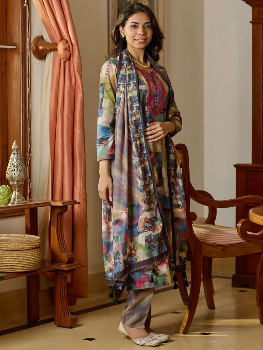 A woman wearing a floral digitally printed cotton suit set with a kurta, pants, and a dupatta, standing in a room with wooden furniture.