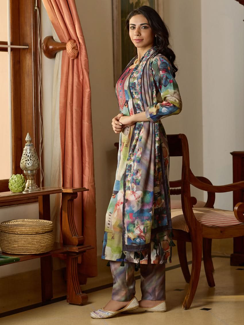A woman wearing a floral digitally printed cotton suit set with a kurta, pants, and a dupatta, standing in a room with wooden furniture.