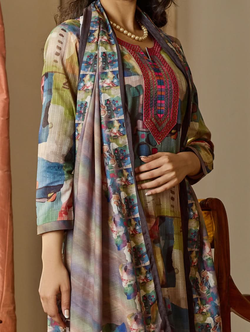 A woman wearing a floral digitally printed cotton suit set with a kurta, pants, and a dupatta, standing in a room with wooden furniture.