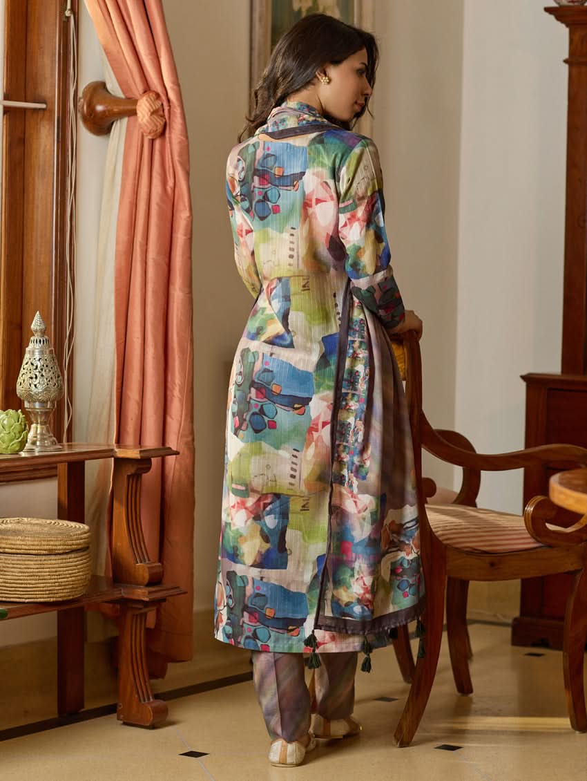 A woman wearing a floral digitally printed cotton suit set with a kurta, pants, and a dupatta, standing in a room with wooden furniture.