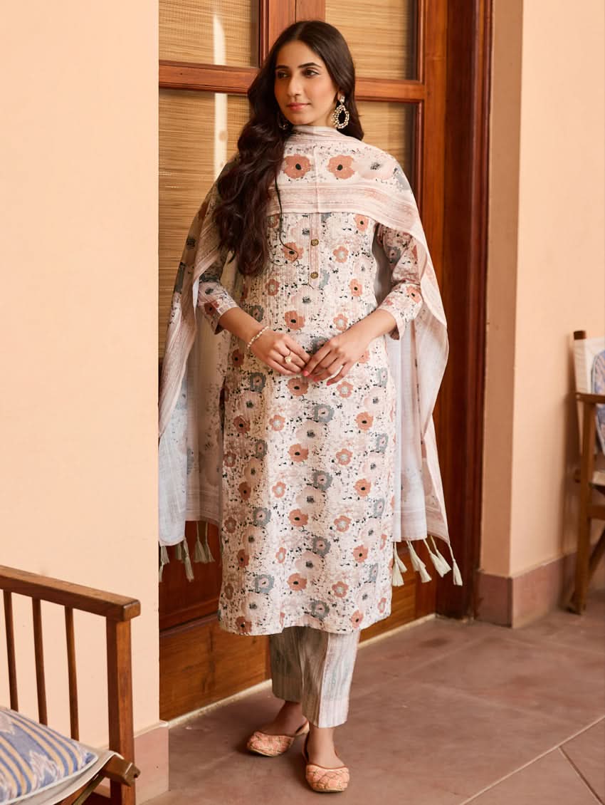 A woman standing in a doorway wearing a traditional white cotton linen suit with floral embroidery, a dupatta over her shoulders, and sandals on her feet.