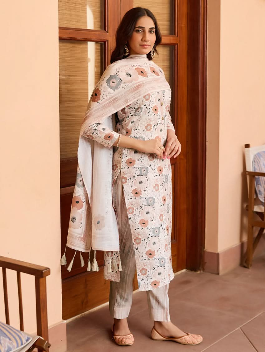 A woman standing in a doorway wearing a traditional white cotton linen suit with floral embroidery, a dupatta over her shoulders, and sandals on her feet.