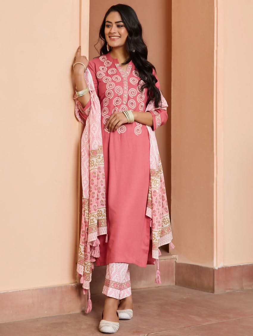 A woman wearing a pink embroidered rayon suit set with a kurta, pants, and a dupatta, standing in a doorway.